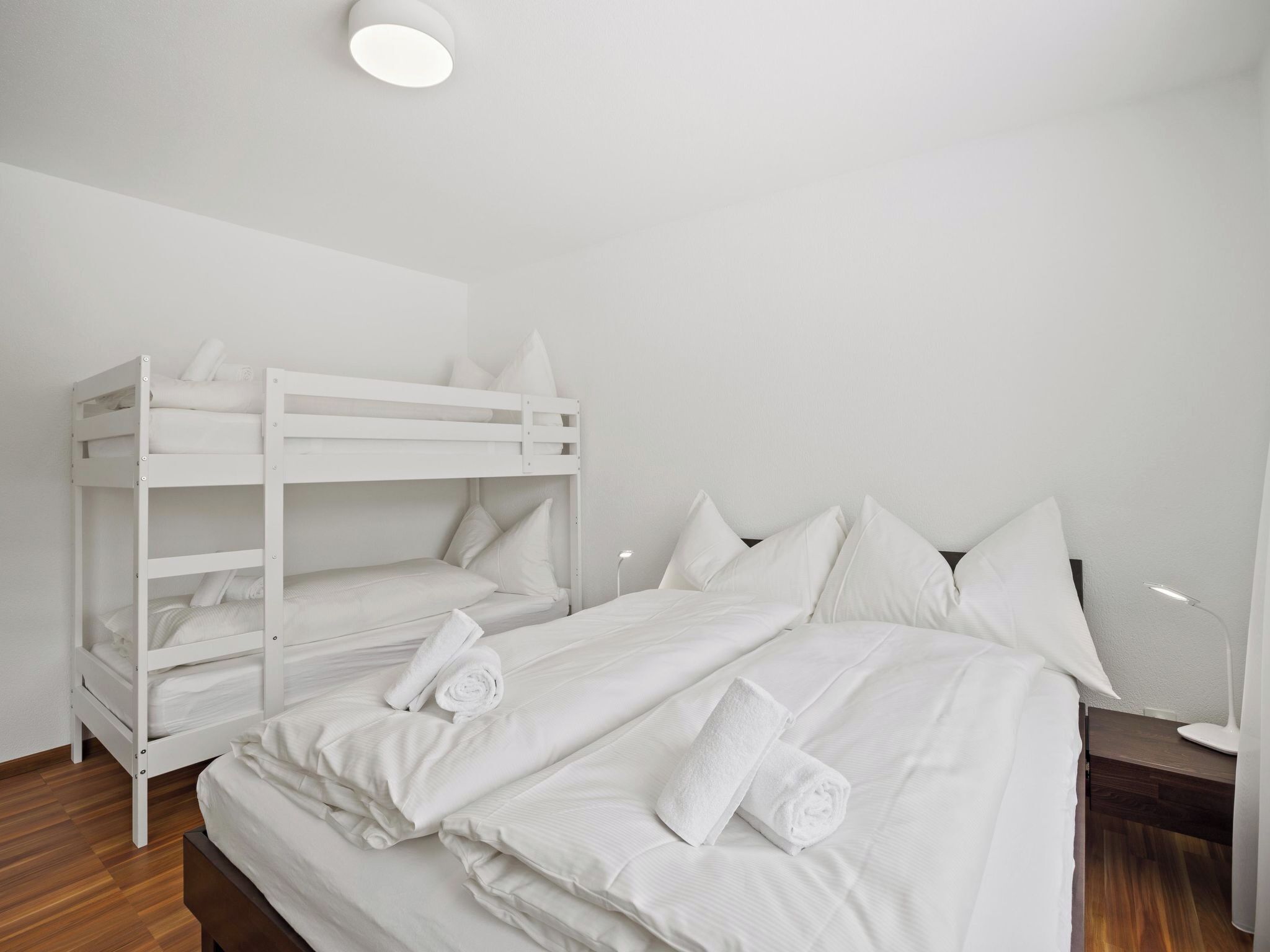 Een slaapkamer met een stapelbed, witte beddengoed en kussens, een nachtkastje en een lamp.