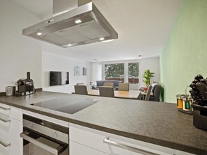 A modern kitchen with a built-in stove, a dining area with a table and chairs, and a view of the outdoors through large windows. - Nr.14 Haus Astoria Appartement/Fewo Nr.14 Haus Astoria by Interhome (Lenzerheide)