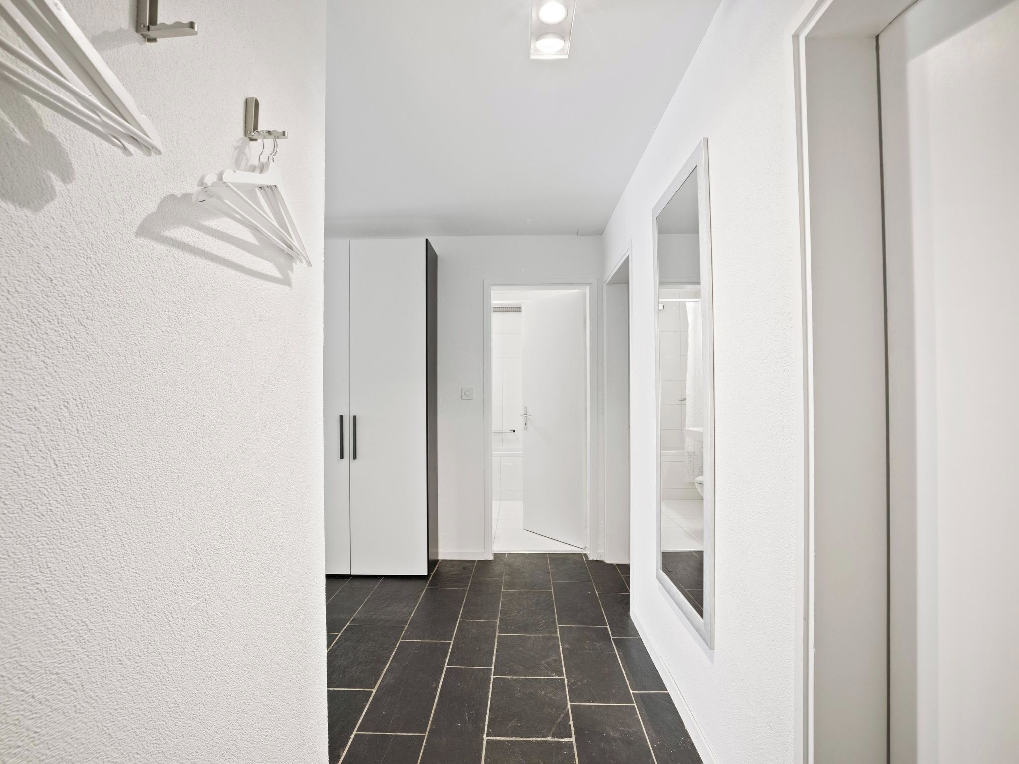 A hallway with white walls, a tiled floor, and a door leading to a bathroom.