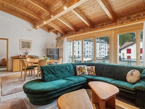 A cozy living area with a green sectional sofa, wooden coffee table, and a dining table with chairs. Large windows offer a view of the cityscape. - Casa Hermi Appartement/Fewo 2.5 Zimmerwohnung by Interhome (Churwalden)