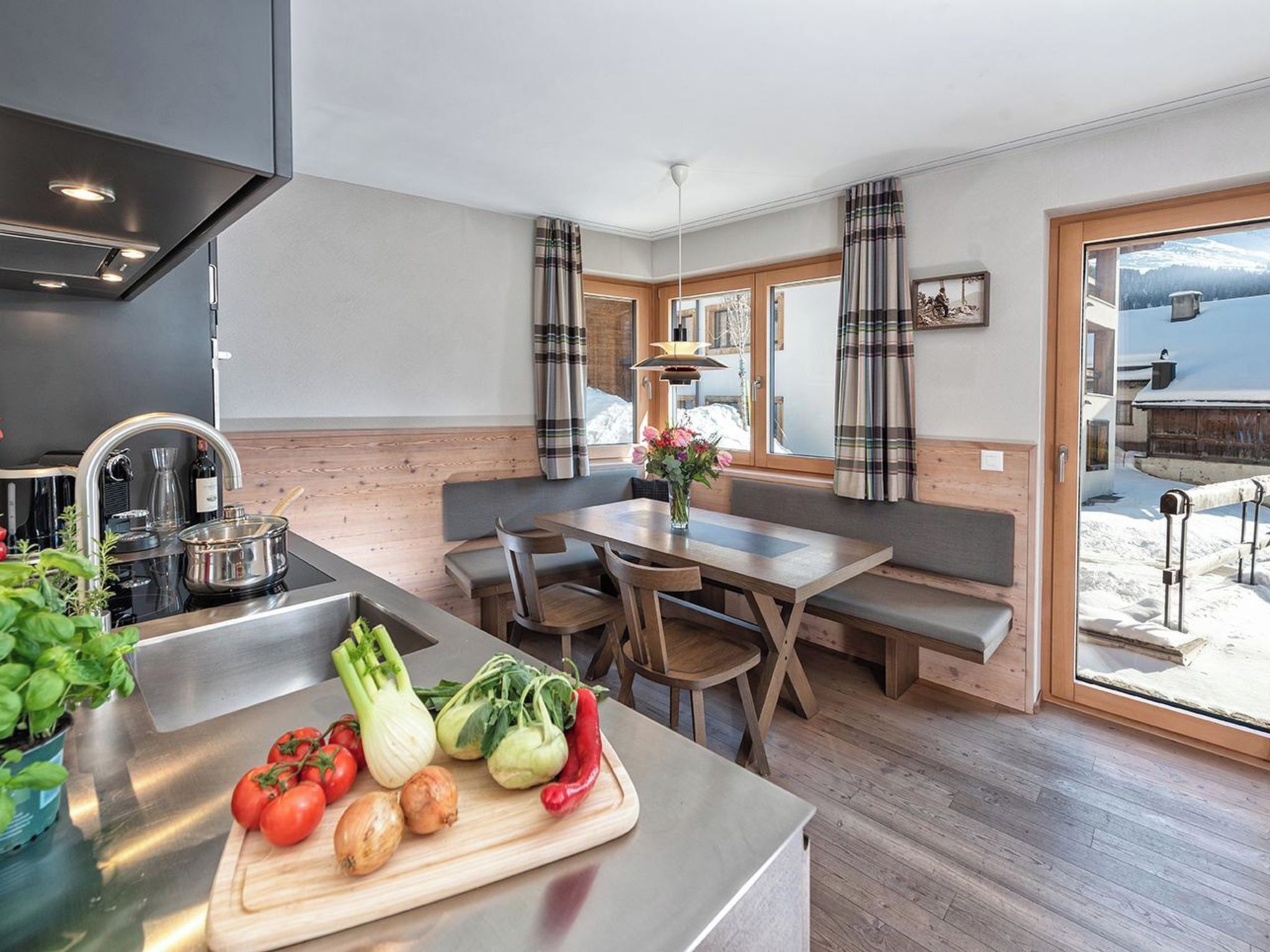 Una cocina moderna con una mesa y banco de madera, vista a un paisaje nevado a través de amplios ventanales, y una variedad de verduras frescas en la encimera.