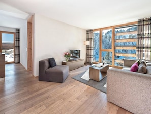 Ein modernes Wohnzimmer mit einer Couch, einem Couchtisch und einem Fernseher. Ein Fenster bietet Blick auf Bäume und eine verschneite Landschaft.