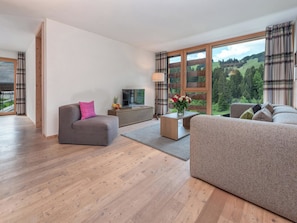 Ein modernes Wohnzimmer mit einer Couch, einem Couchtisch und einem Fernseher. Ein großes Fenster mit Vorhängen bietet Blick auf eine bewaldete Gegend.