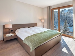 Ein Schlafzimmer mit einem großen Bett, einem Nachttisch, einer Lampe und einem Fenster mit Blick auf eine verschneite Landschaft.