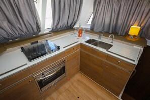 A compact kitchen with a sink, stove, and oven, wooden cabinets, and a window with curtains. - Sun-Kissed Sailing Adventure in Split, Croatia (Kaštel Gomilica)