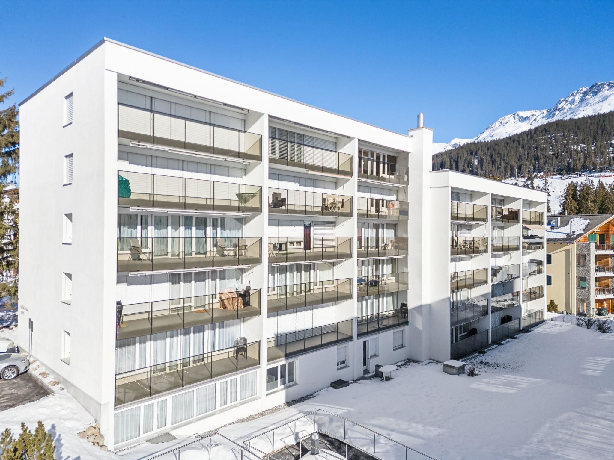 A multi-story hotel with balconies, a parking area, and a snowy mountain backdrop.