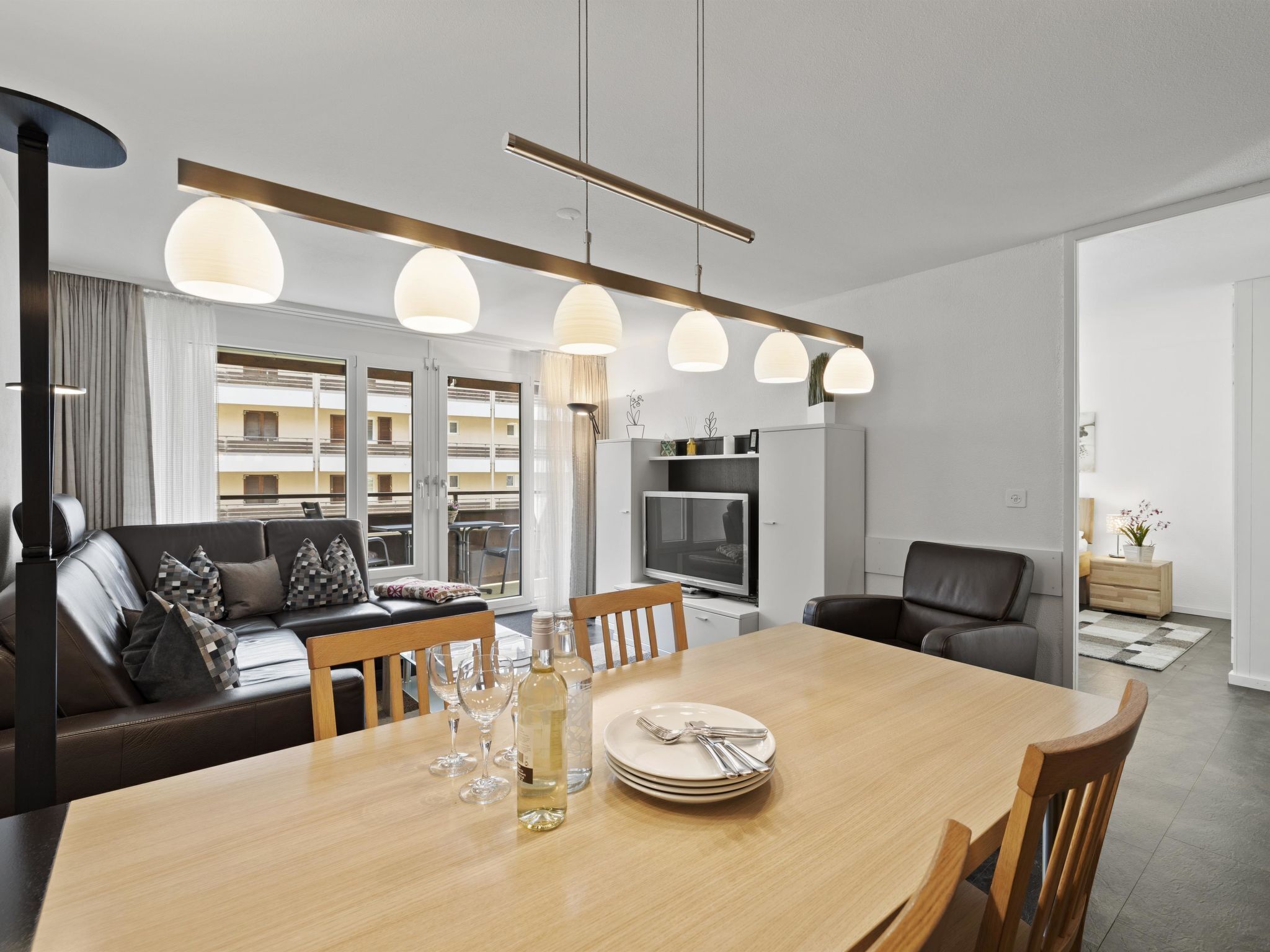 Ein modernes Wohnzimmer mit Essbereich, ausgestattet mit einer Couch, einem Esstisch gedeckt mit Tellern, Gläsern und einer Flasche, sowie Pendelleuchten darüber.