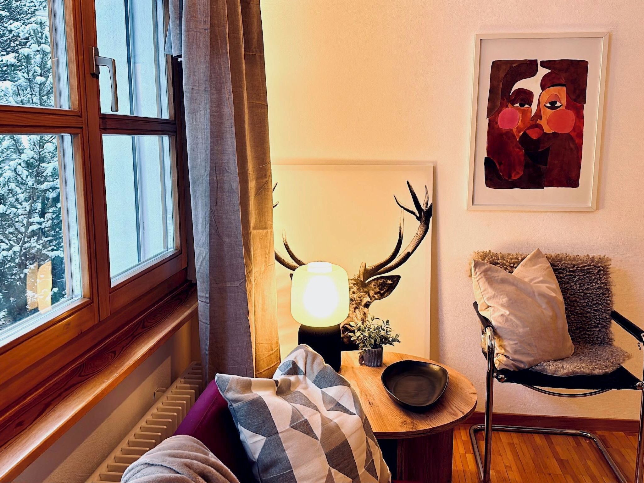 Ett mysigt rum med ett fönster, en lampa med renhorn, en stol och en tavla på väggen.