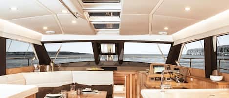 Interior de un yate con comedor, asientos, cocina y vistas al mar.