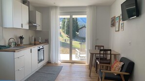 Una cocina moderna con pileta, estufa y placares, un comedor con mesa y sillas, y un amplio ventanal corredero que da a un balcón con vista a una casa y árboles.