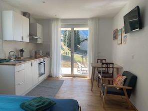 A modern kitchen with a sink, stove, and cabinets, a dining area with a table and chairs, and a large sliding glass door leading to a balcony with a view of a house and trees. - Studio Bossihang Appartement/Fewo, Dusche oder Bad, WC, 1 Schlafraum by Interhome (Lenzerheide)