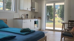 Un dormitorio moderno con una cama grande, un banco de madera, una kitchenette y un ventanal corredero que da a un balcón.