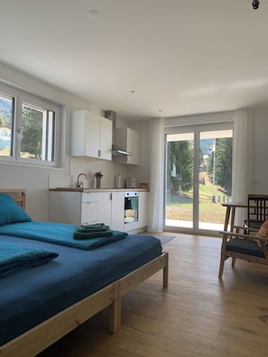 A modern bedroom with a large bed, a wooden bench, a kitchenette, and a sliding glass door leading to a balcony. - Studio Bossihang Appartement/Fewo, Dusche oder Bad, WC, 1 Schlafraum by Interhome (Lenzerheide)