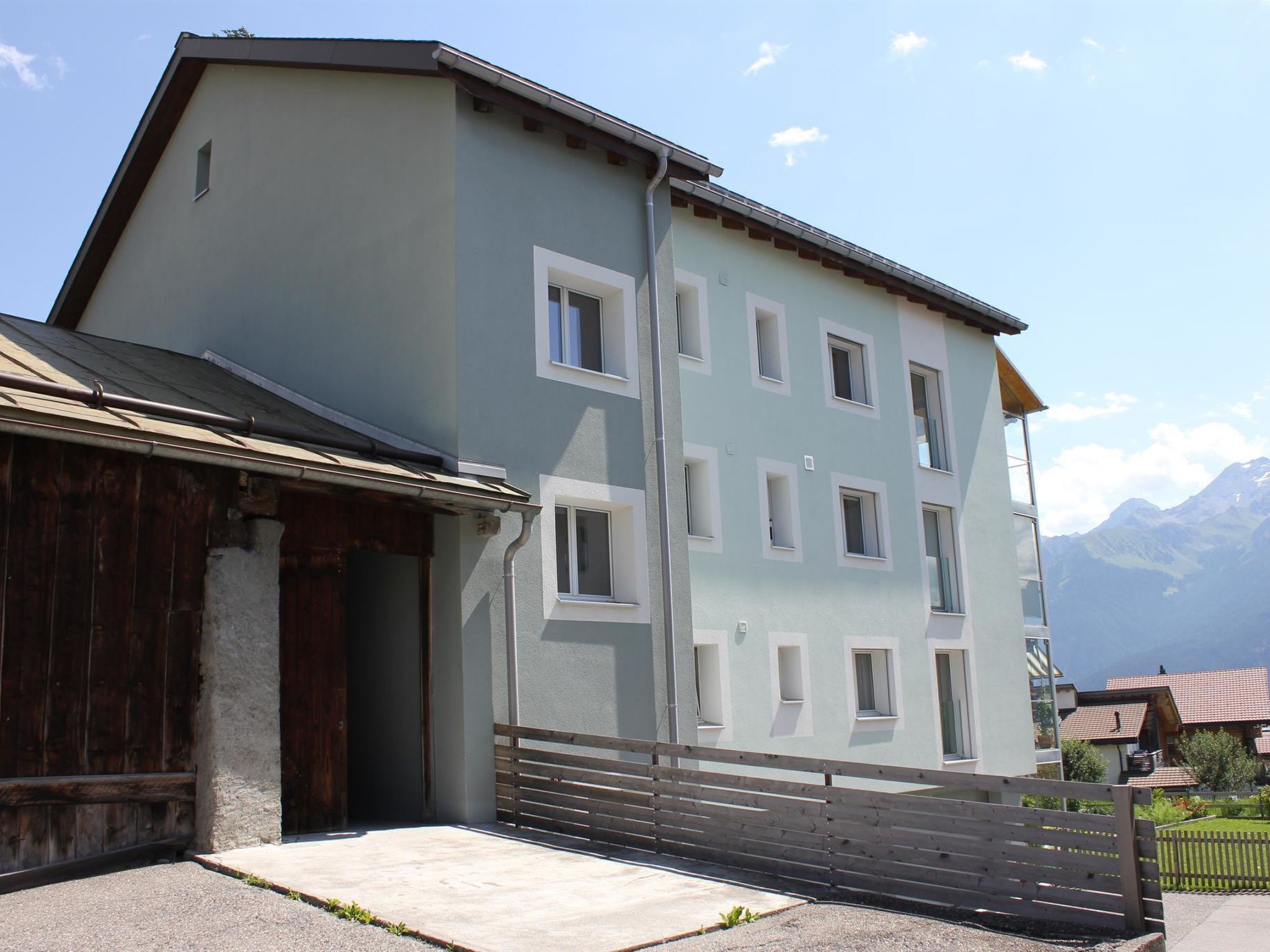 A multi-story building with a light facade, a wooden structure to the left, and a clear view of mountains in the background.