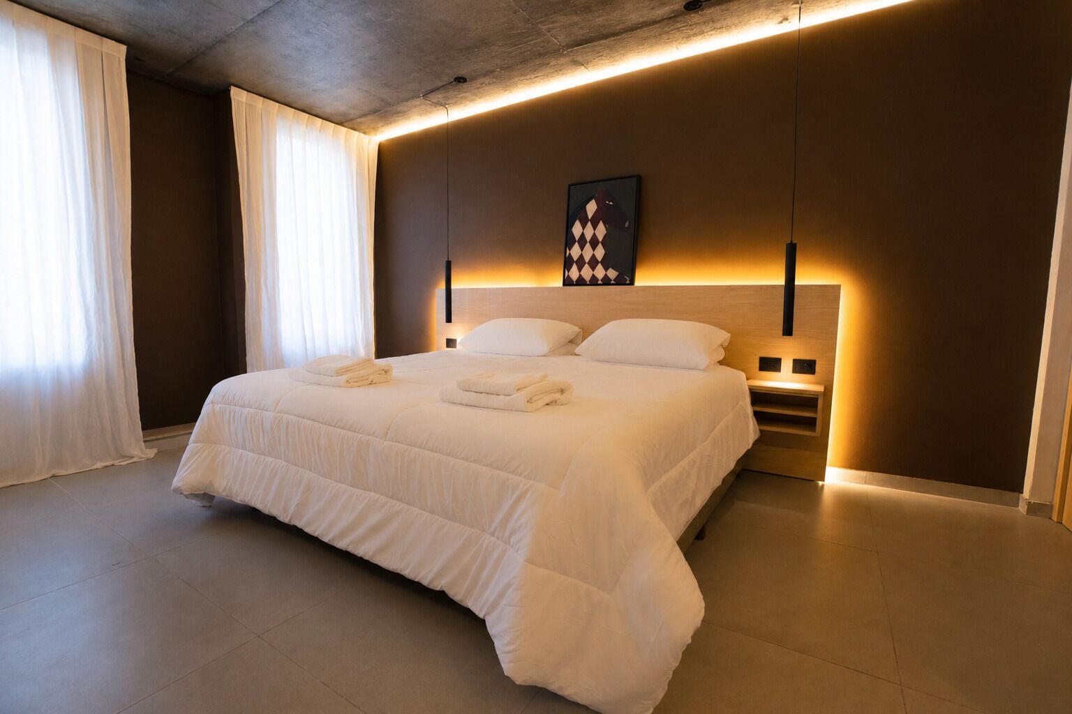 A hotel room with a large bed, white linens, a headboard with integrated lighting, and a framed artwork above the bed.