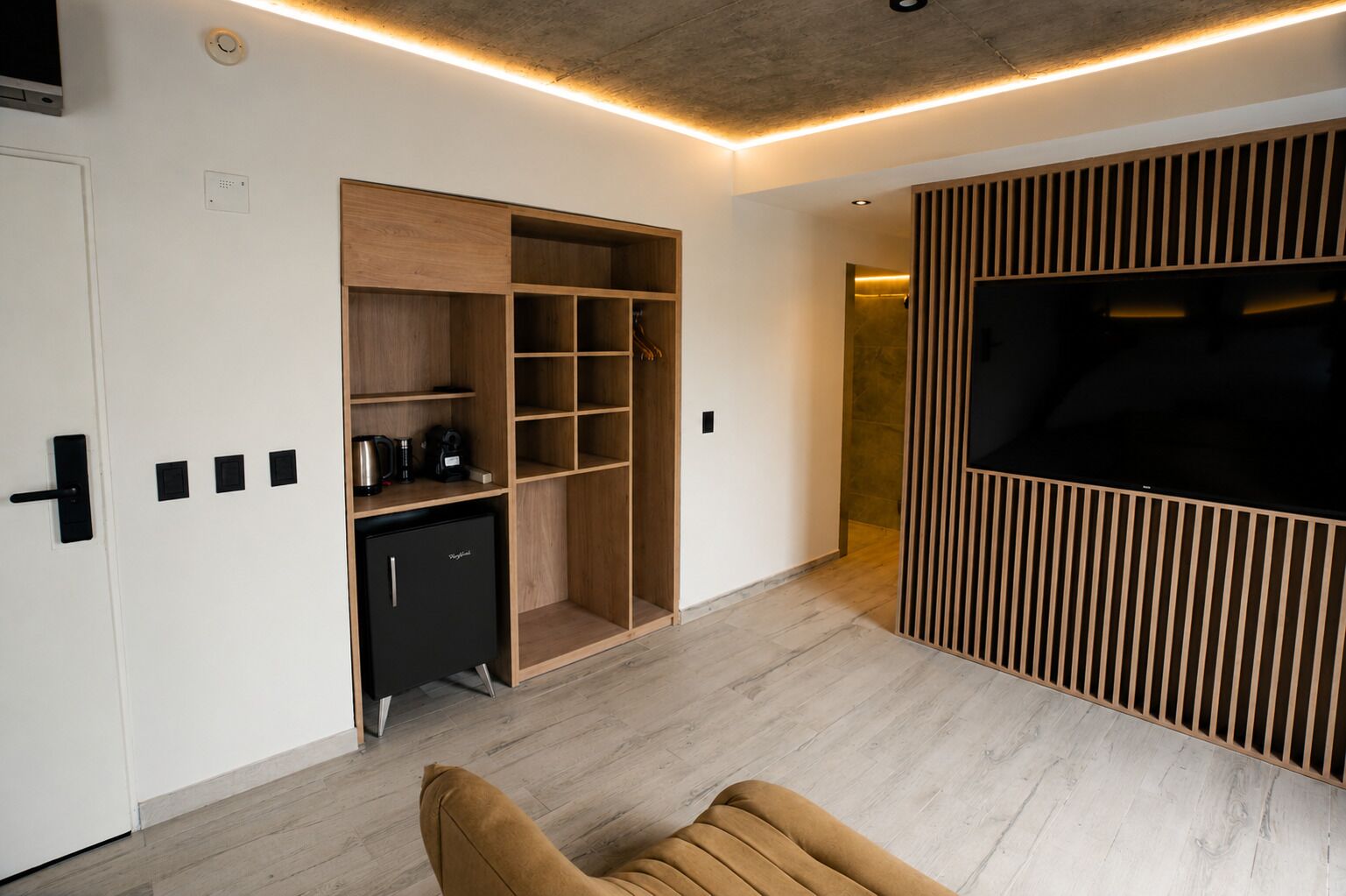 Una sala de estar moderna con televisor empotrado, estantería de madera y una pequeña cocina.