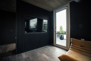 Una sala moderna con un televisor de pantalla plana grande montado en una pared oscura, una puerta corrediza de vidrio que da a un espacio exterior, y un sofá marrón.