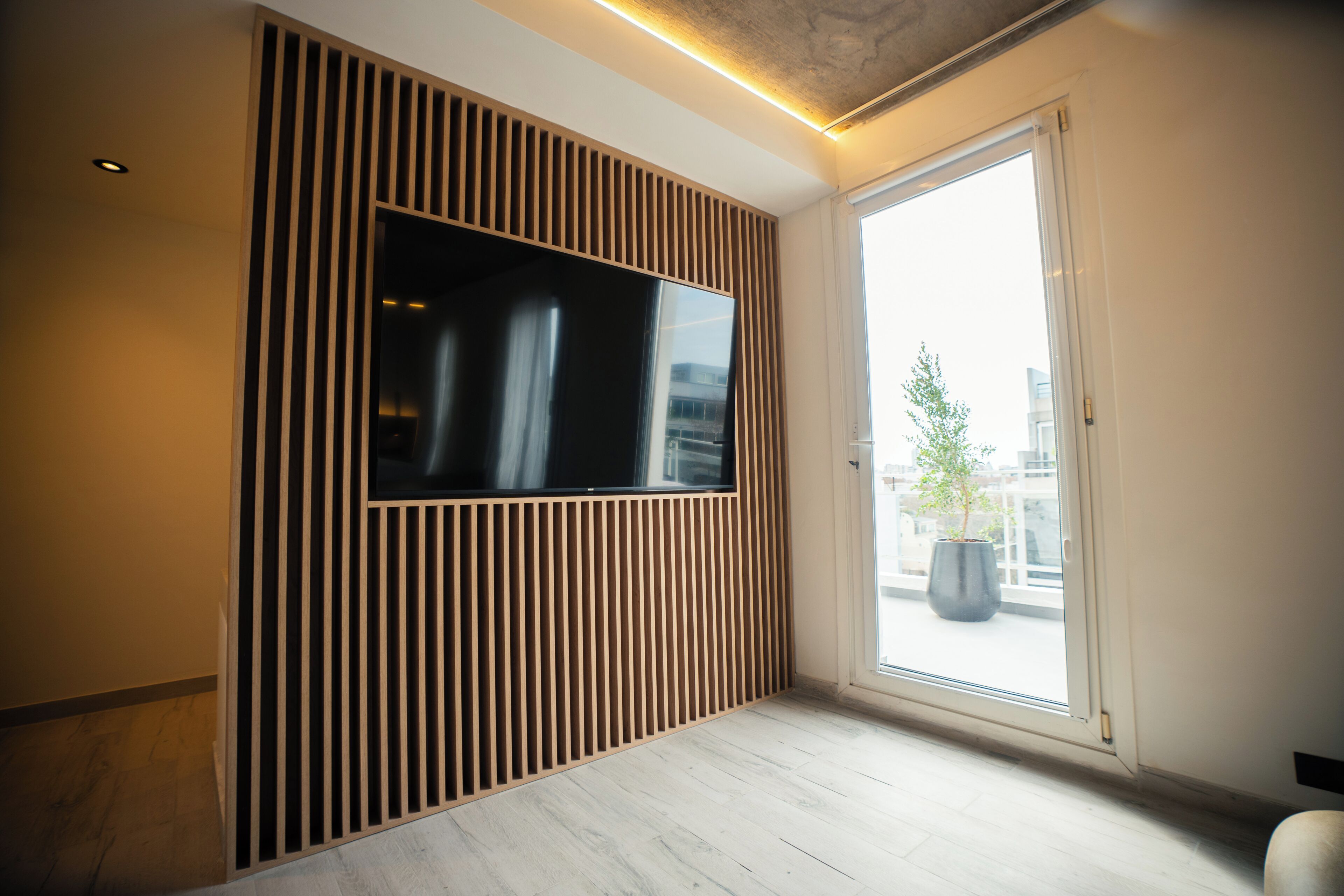 Un interior moderno con un televisor de pantalla plana montado en una pared de paneles de madera, un ventanal con vistas a edificios y una planta en el alféizar.