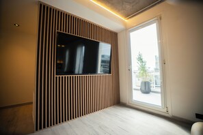 Un interior moderno con un televisor de pantalla plana montado en una pared de paneles de madera, un ventanal con vistas a edificios y una planta en el alféizar.