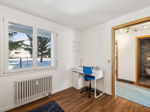 A room with a white desk, a blue chair, a radiator, and a window with a view of trees and buildings. - Nr.06 Haus Sursilvana Appartement/Fewo,   Nr.06 Haus Sursilvana by Interhome (Lenzerheide)