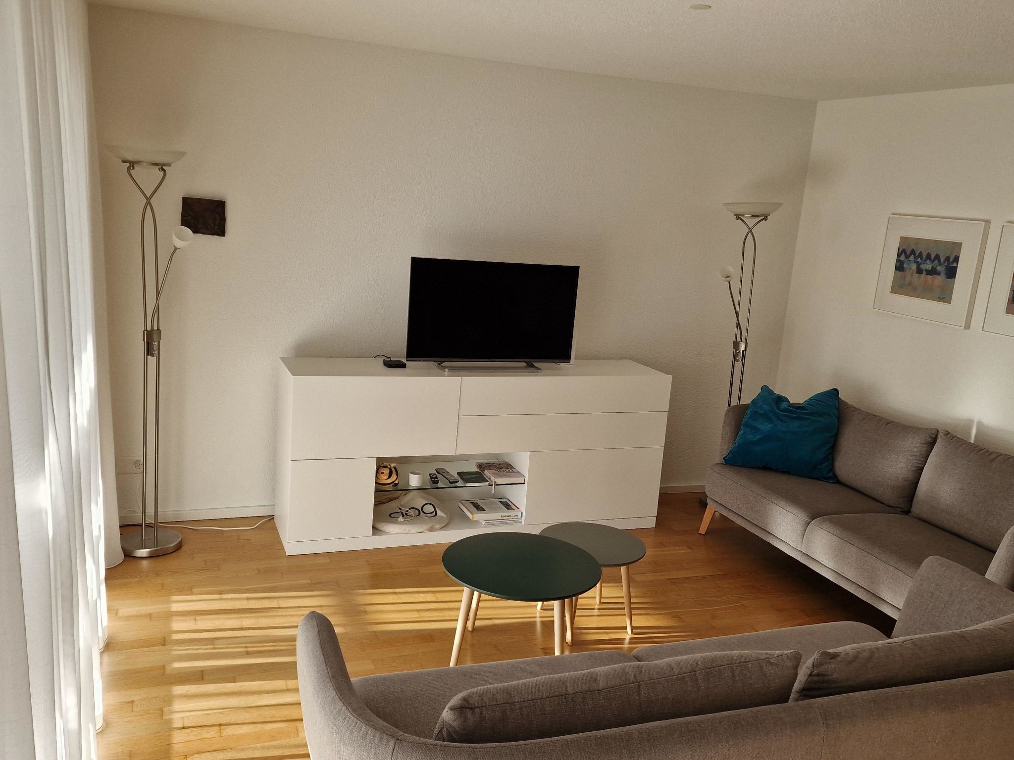 Un salon avec un téléviseur à écran plat posé sur un meuble blanc, un canapé gris, une petite table ronde et un lampadaire.