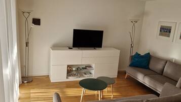Un salon avec un téléviseur à écran plat posé sur un meuble blanc, un canapé gris, une petite table ronde et un lampadaire.