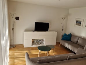 Ein Wohnzimmer mit Flachbildfernseher auf einer weißen Kommode, einem grauen Sofa, einem kleinen runden Tisch und einer Stehlampe.