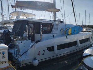 A boat named 'BALI 42' docked at a marina with other boats, a cabin with windows, and a blue lifebuoy on deck.