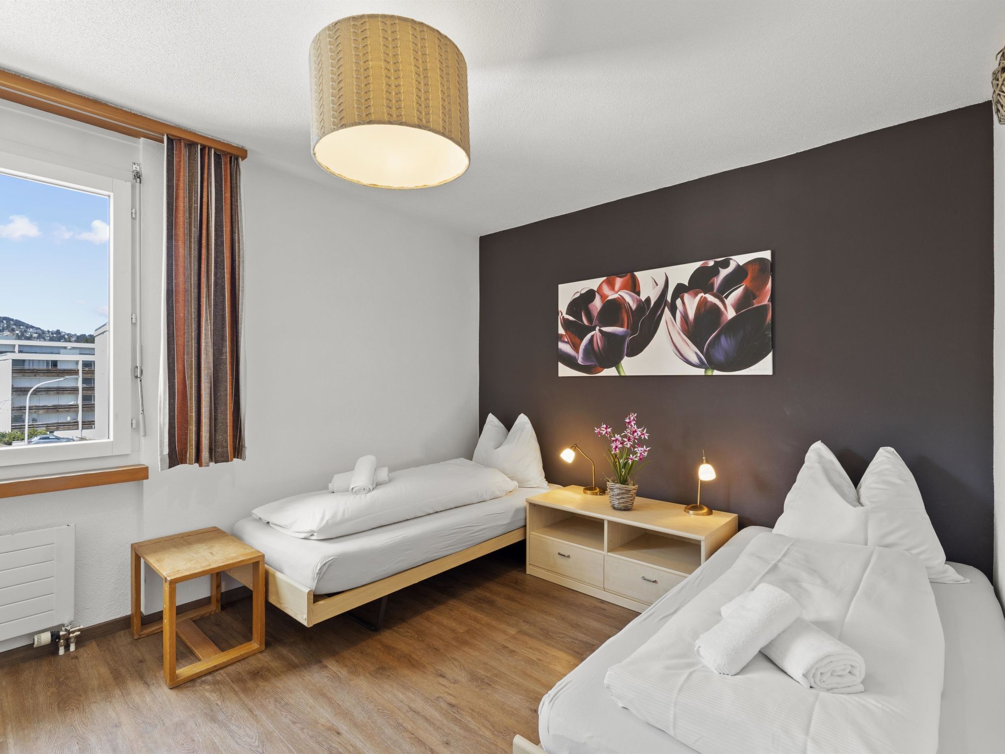 A modern bedroom with a bed, a sofa, a bedside table with lamps, and a window with curtains.