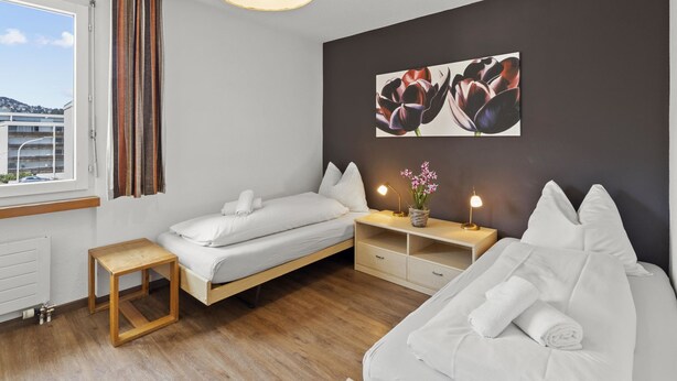 A modern bedroom with a bed, a sofa, a bedside table with lamps, and a window with curtains.