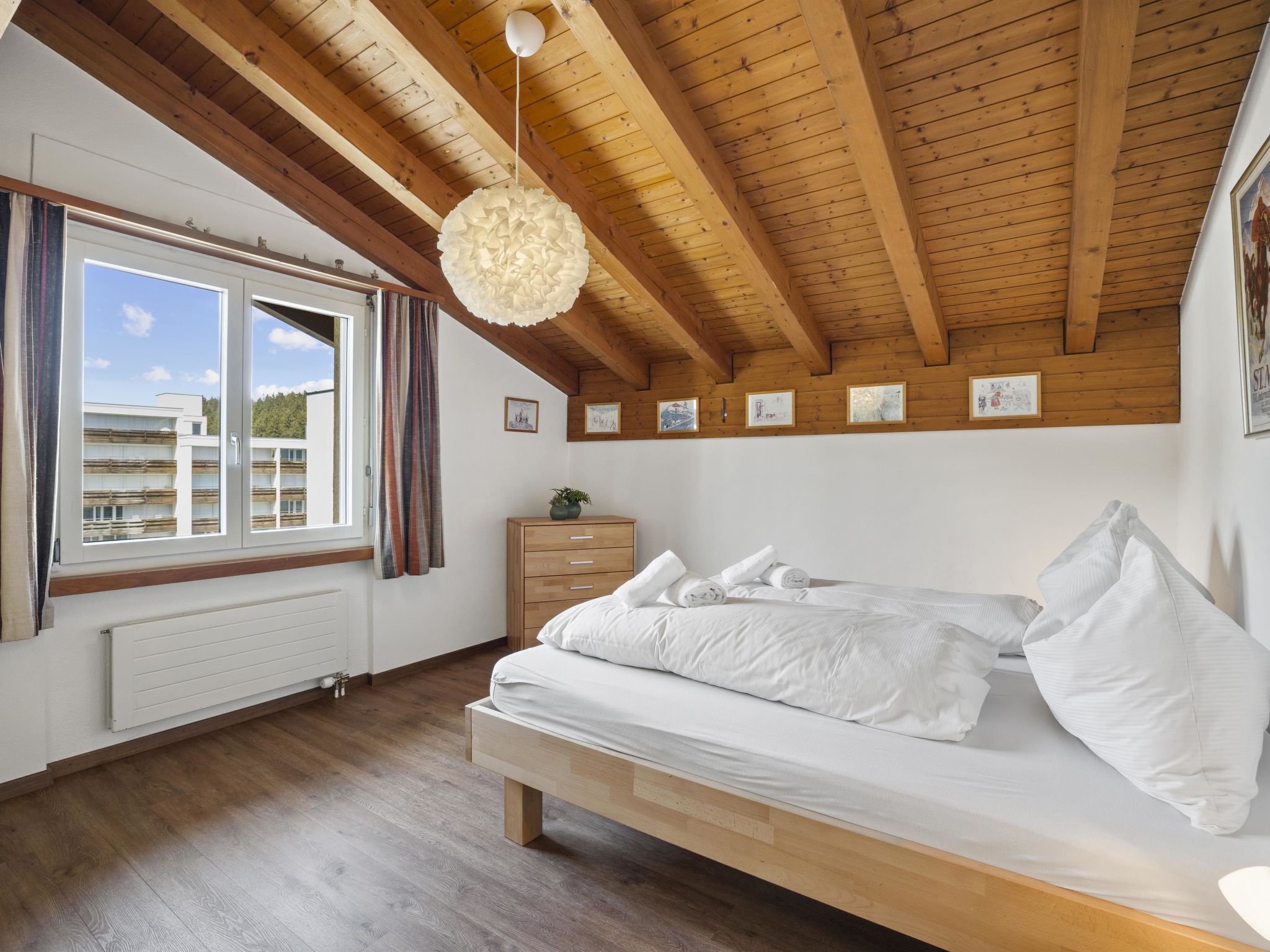 A bedroom with a wooden ceiling, a bed with white bedding, a window with curtains, and a radiator.