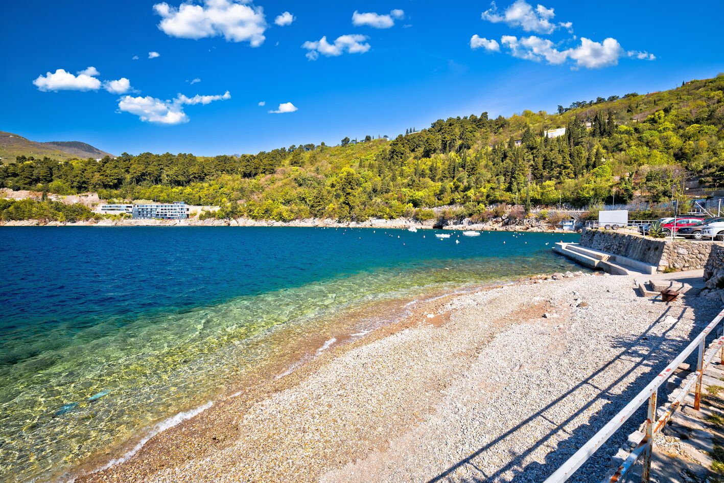 A coastal beach with clear water, a rocky shoreline, and a building with a balcony overlooking the sea.