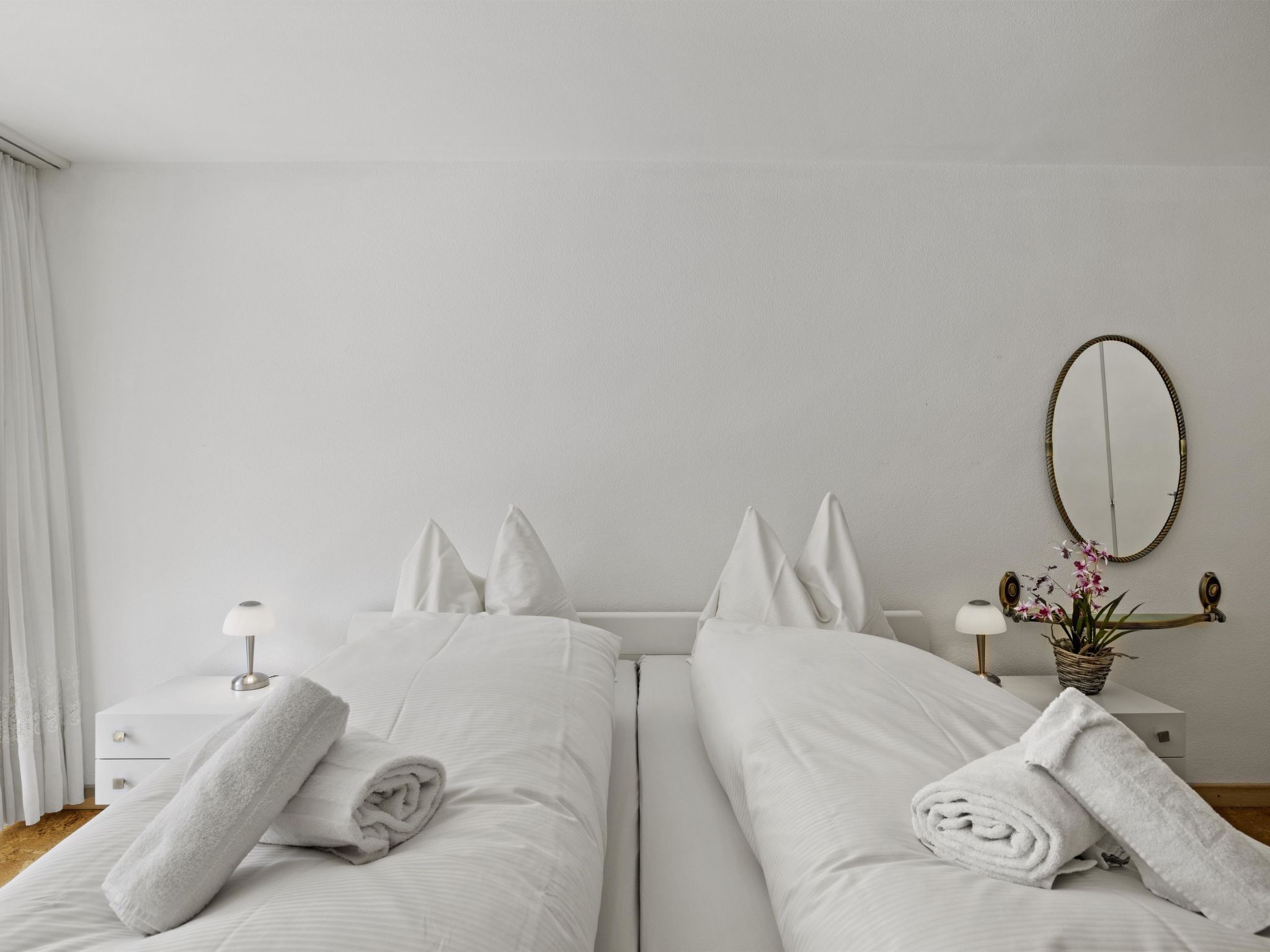 Habitación de hotel con dos camas, ropa de cama blanca, una mesita de noche con lámpara, un espejo y una orquídea en maceta.