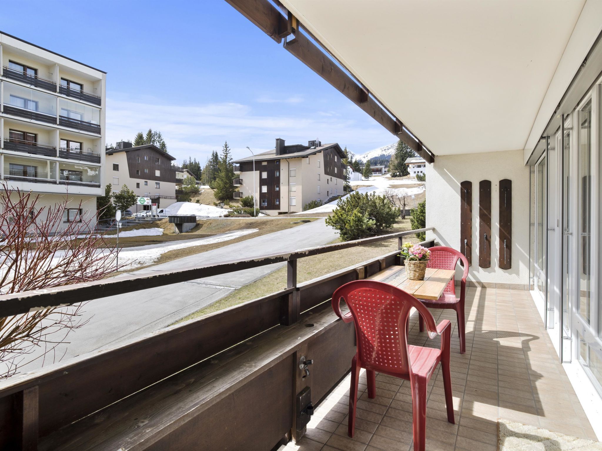 A balcony with a table and chairs, overlooking a residential area with buildings and a snowy landscape.
