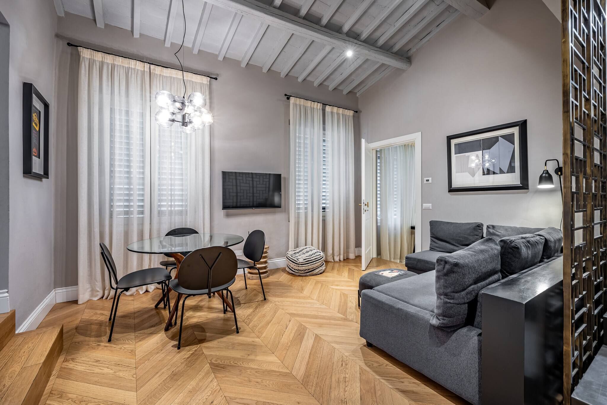 A modern living room with a herringbone wood floor, a grey sofa, a dining table with chairs, and a chandelier.