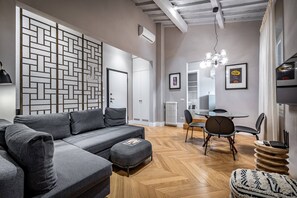 Ein modernes Wohnzimmer mit einem grauen Ecksofa, einer Wand mit Fischgrätmuster, einem Essbereich mit rundem Tisch und Stühlen sowie einer sichtbaren Klimaanlage.