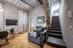 Ein modernes Wohnzimmer mit grauem Ecksofa, Holzboden im Fischgrätmuster, einem Flachbildfernseher und einer Treppe, die in eine obere Ebene führt.