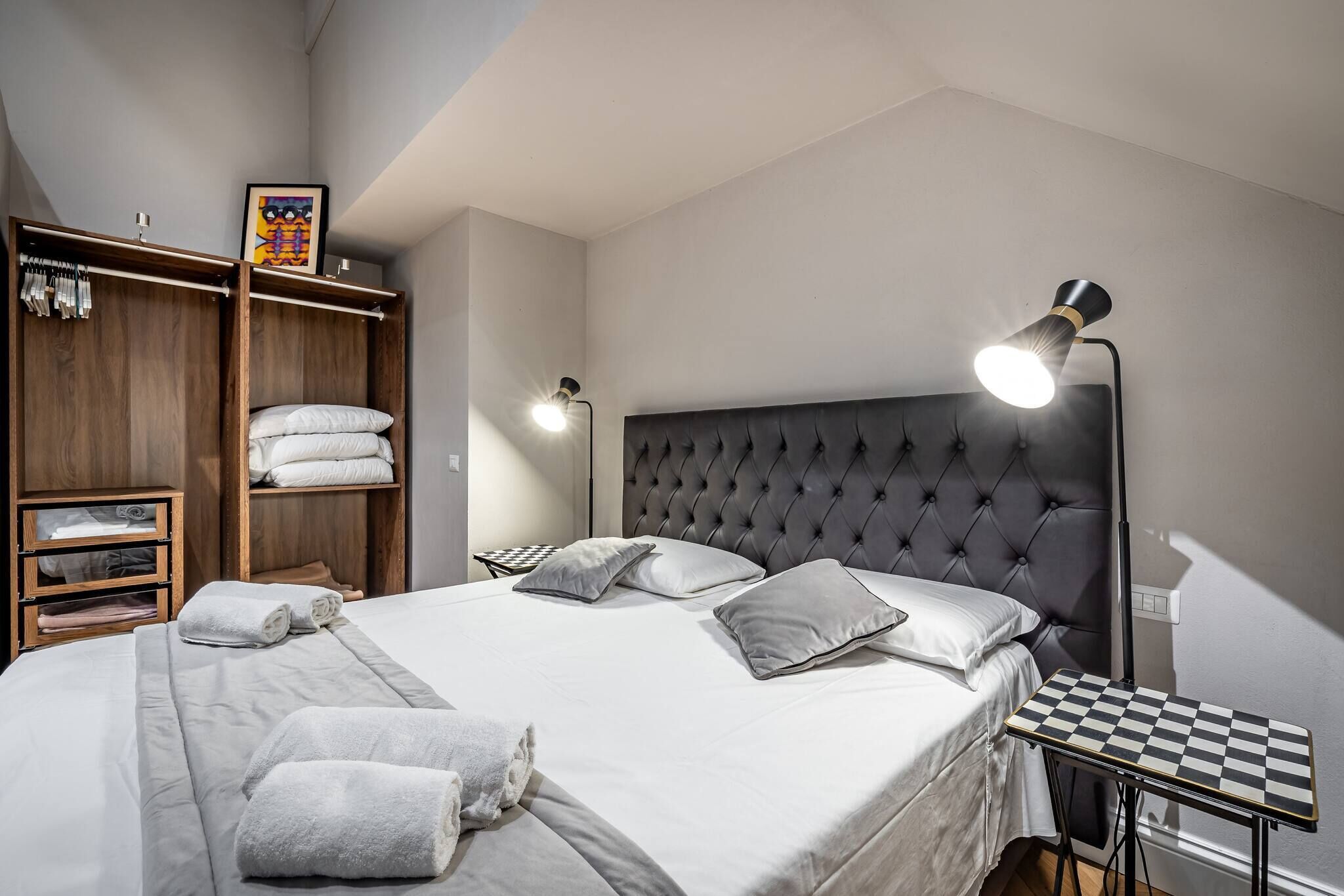 Una cama bien hecha con almohadas y toallas, una mesita de noche con lámpara, y un armario con ropa colgada.