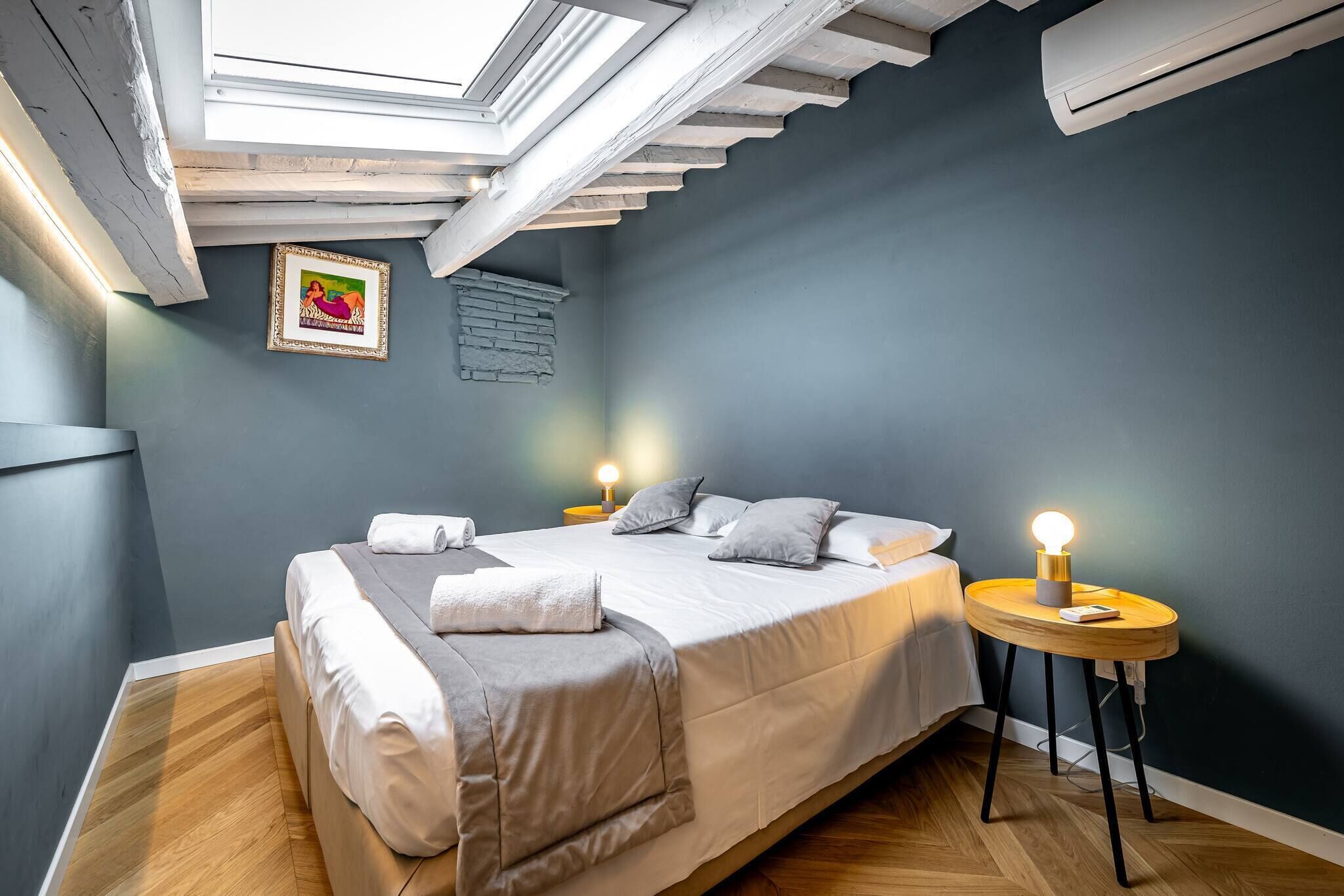 A bedroom with a bed, bedside tables, a wall-mounted picture, and a skylight.