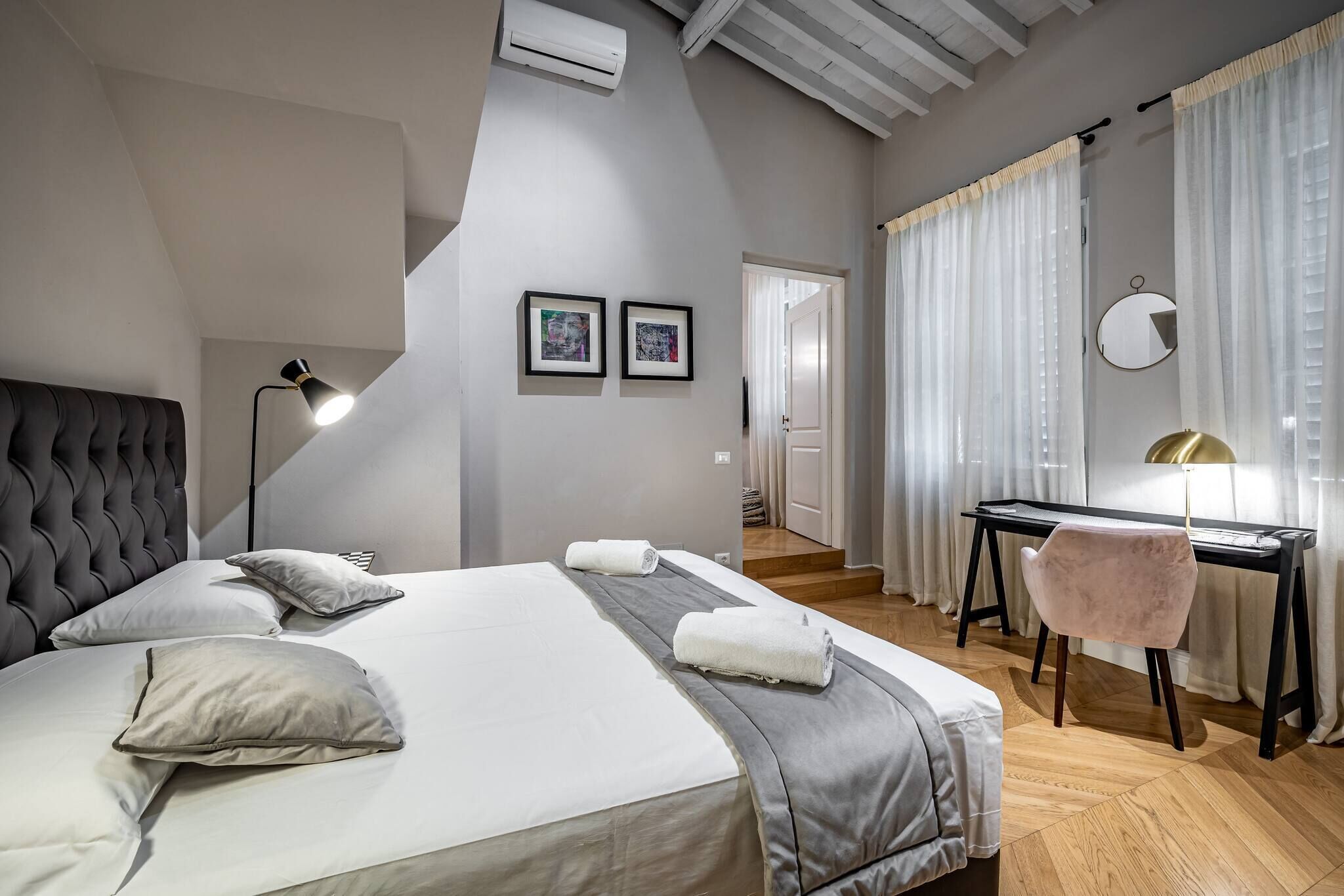 A modern bedroom with a large bed, a desk, and a chair. There are two framed pictures on the wall, a mirror, and a ceiling fan.