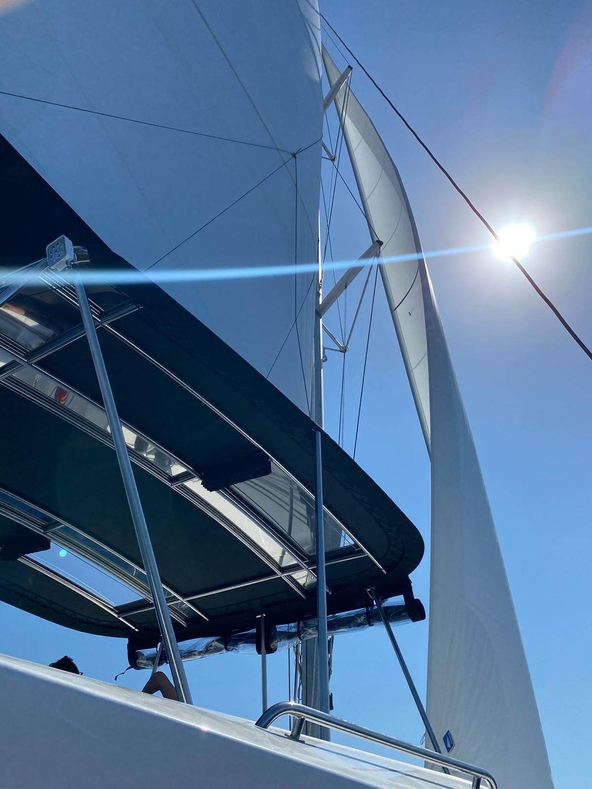 Een zeilboot met een zichtbare mast en zeilen, een heldere lucht en een zon die achter de zeilen schijnt.