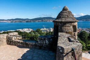 Ein Steingebäude mit einem kegelförmigen Dach, das auf eine Küstenstadt mit freiem Blick auf das Meer und ferne Berge blickt.
