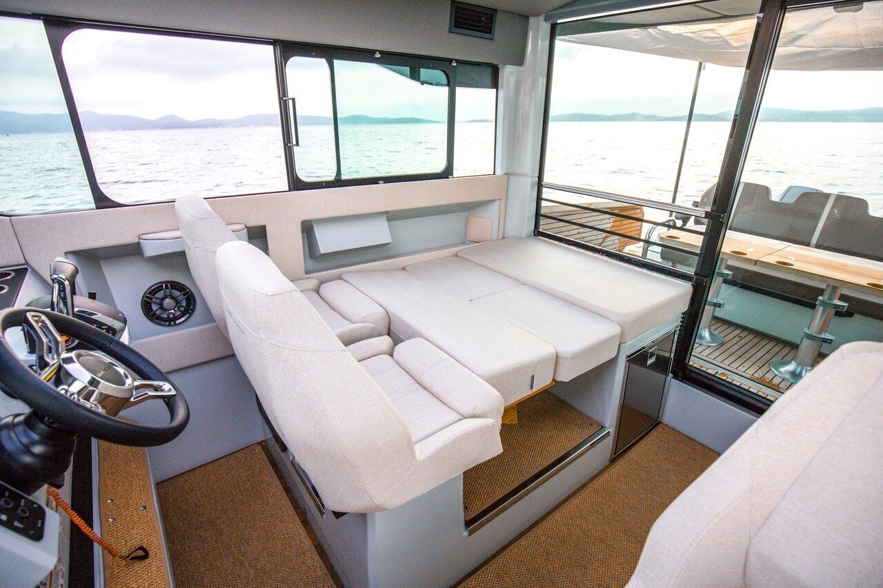 Interior de un barco con timón, panel de control, asientos acolchados y vista al agua.