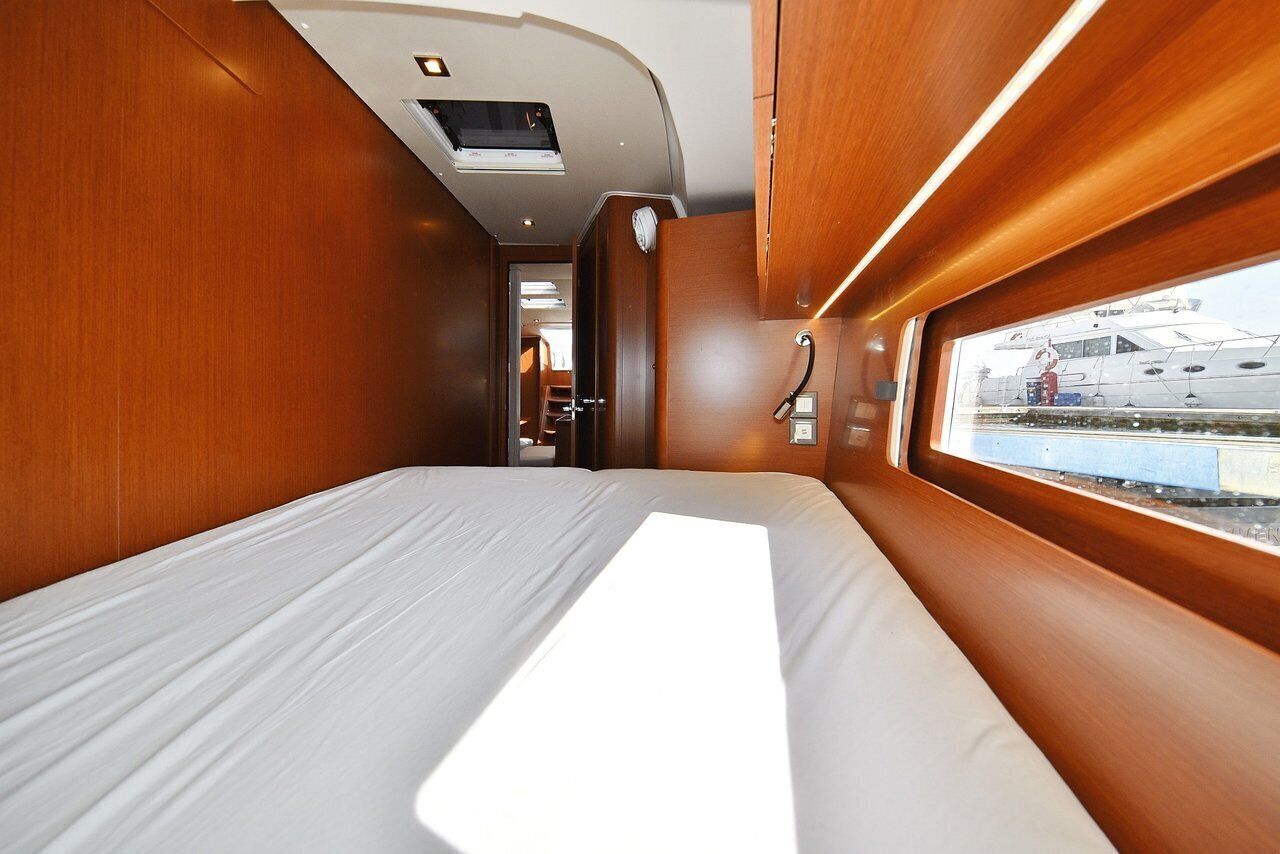 A narrow, well-lit corridor with wooden walls, a bed with white bedding, and a window with a view of a docked boat.