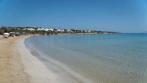 Uma praia com águas cristalinas, areal arenoso e edifícios ao fundo.