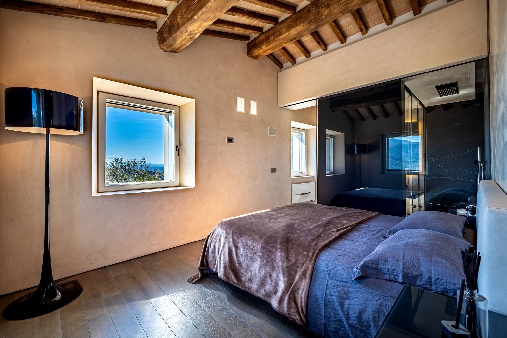 Una camera da letto con un'ampia finestra, un letto con coperta marrone, una lampada da terra e uno specchio.