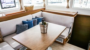 A boat interior with a wooden table, cushioned seating, a window with a view of boats, and a bottle of wine. - Sun-Kissed Sailing Adventure in Sibenik, Croatia (Primošten)