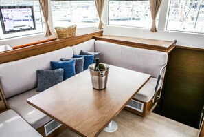 A boat interior with a wooden table, cushioned seating, a window with a view of boats, and a bottle of wine. - Sun-Kissed Sailing Adventure in Sibenik, Croatia (Primošten)