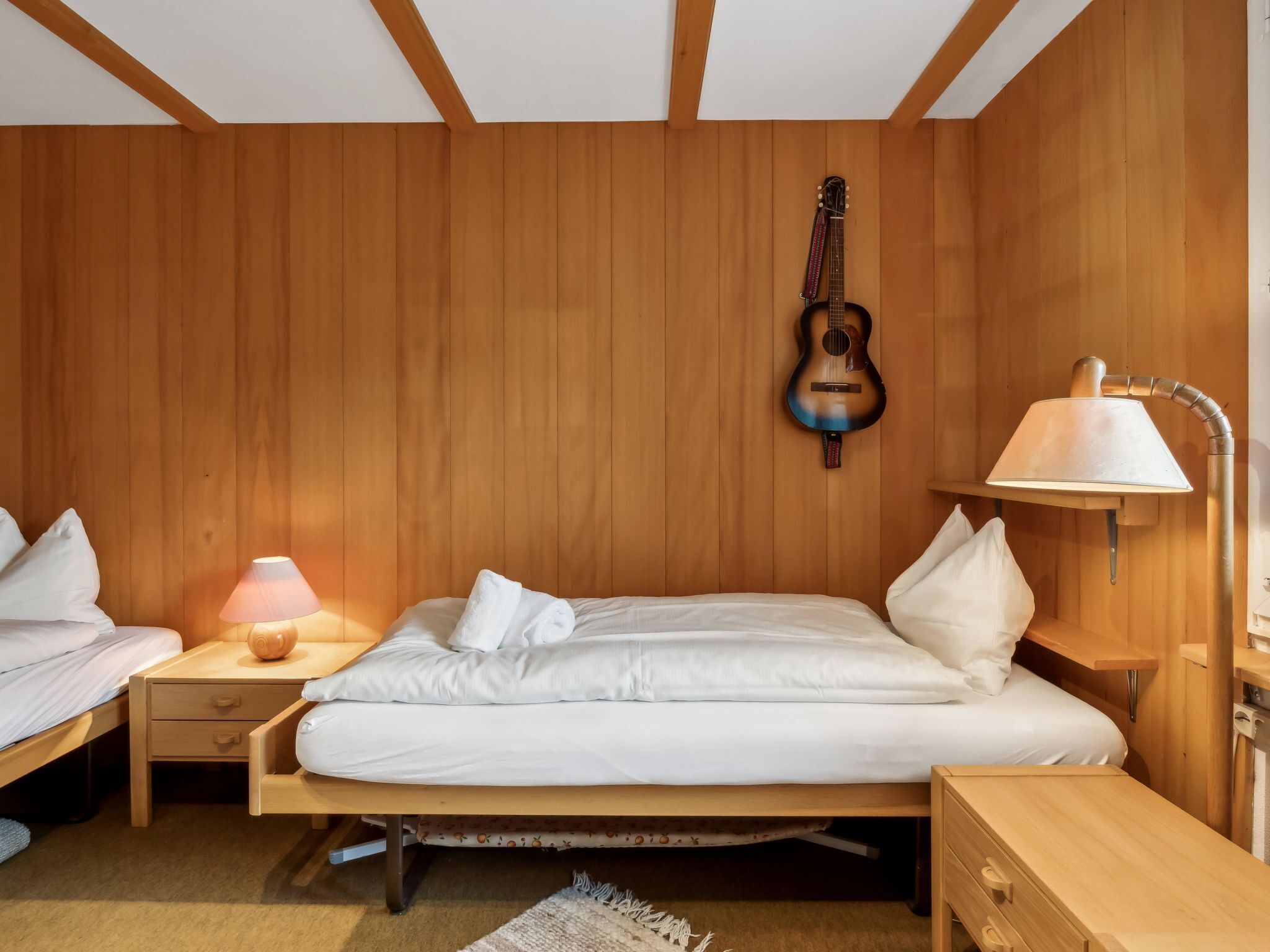 Ein Zimmer mit Holzwänden, einem Bett, einem Nachttisch mit Lampe und einer Gitarre an der Wand.