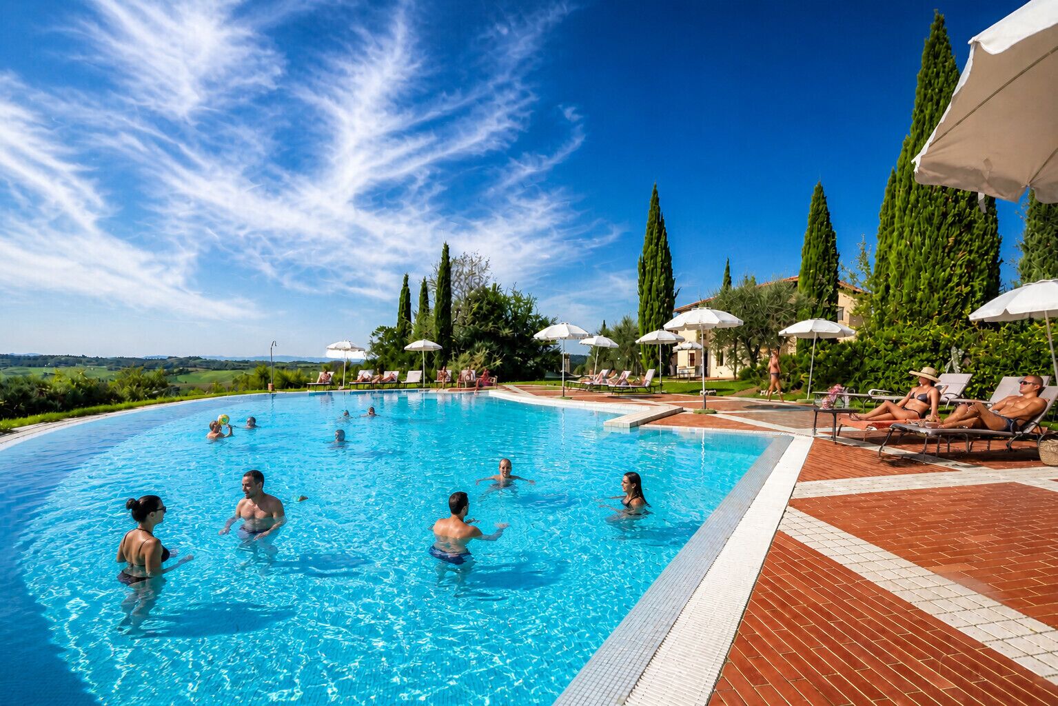 Una piscina con gente, rodeada de sillas de descanso y sombrillas.